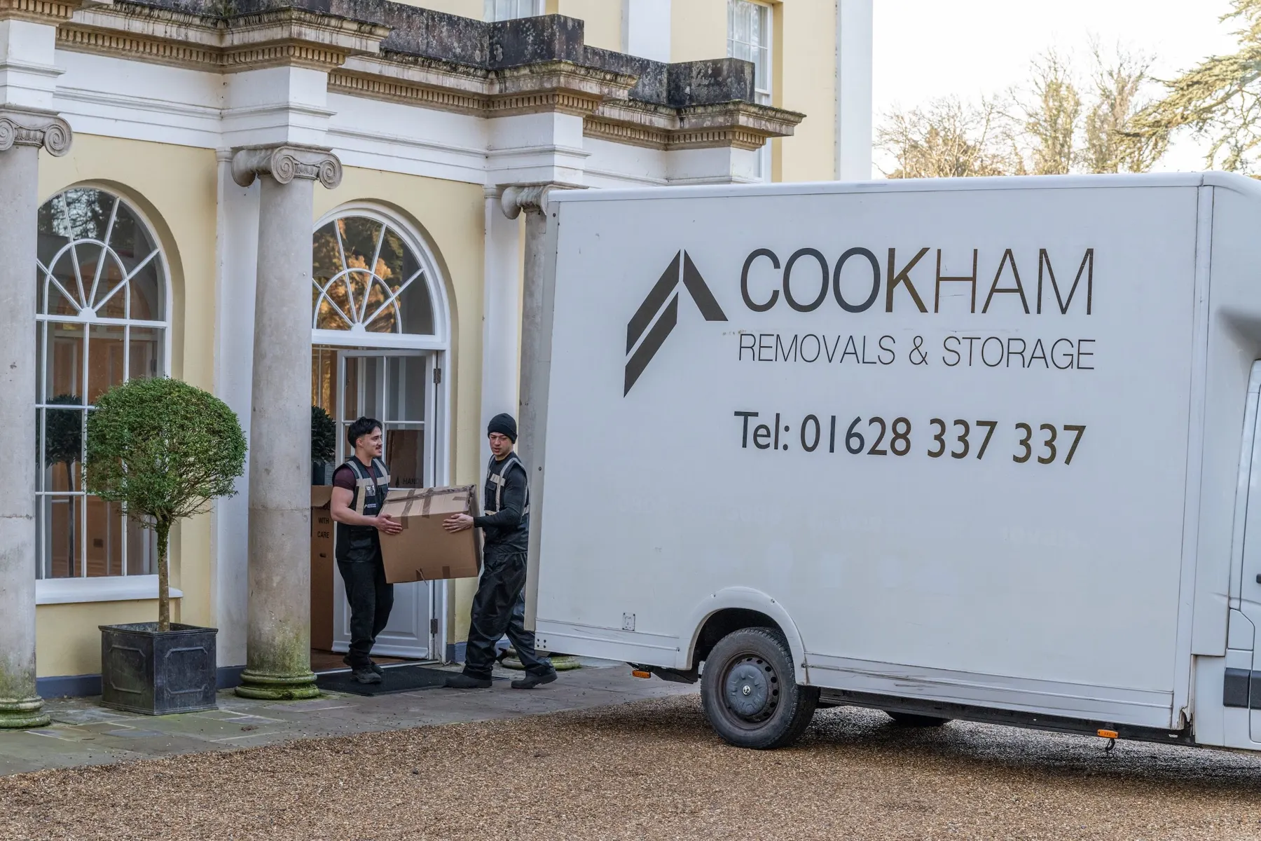 Two Cookham Removals & Storage crew members carrying a box to the Luton van at the pillared entrance of the Georgian house