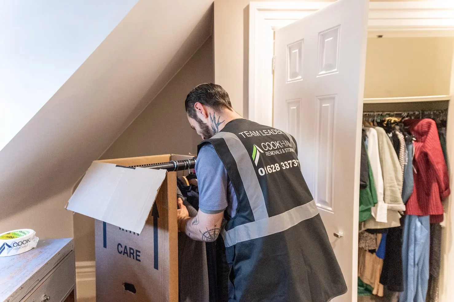A Cookham Removals & Storage crew member packing a box in a loft room with a sloping ceiling and a rail of clothes