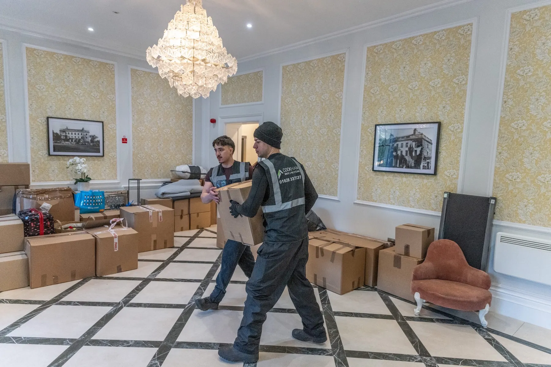 Two Cookham Removals & Storage crew members carrying boxes across a tiled hallway with a chandelier